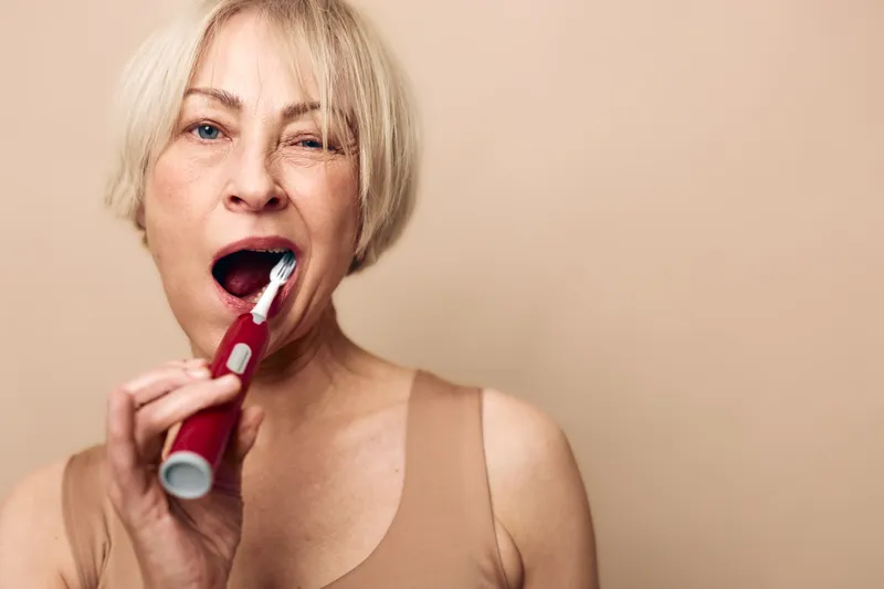 Middle aged woman with short blonde hair brushing teeth with electric toothbrush in beige top. Close-up on neutral background. Concept of oral hygiene, dental care, and daily routine.
