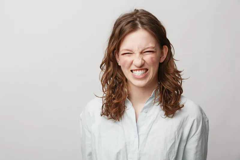 Young woman with curly hair wearing white shirt grimaces with eyes closed and teeth clenched. Studio portrait on light background. Concept of emotion, expression, and candid moment.