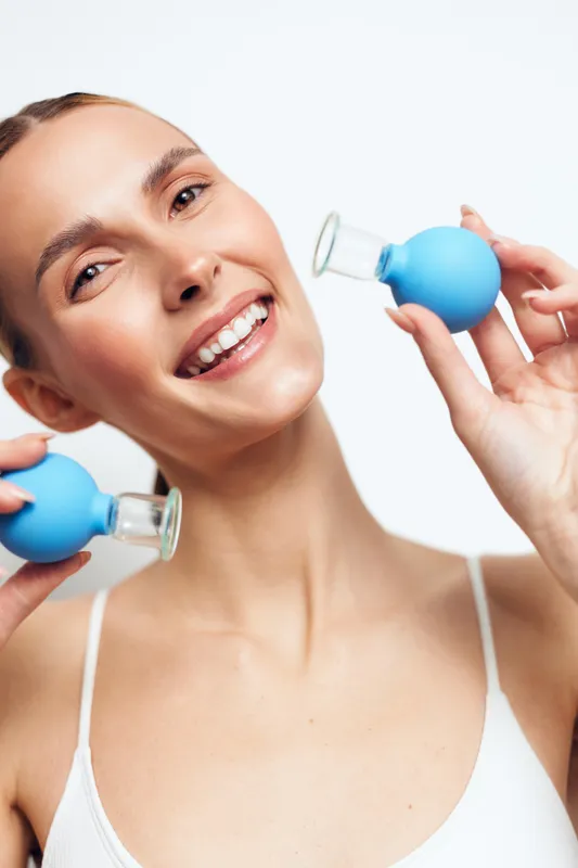 A smiling woman uses facial exercise tools in a bright setting, promoting skincare, health, and wellness. A clear concept of self care and beauty through simple routines.