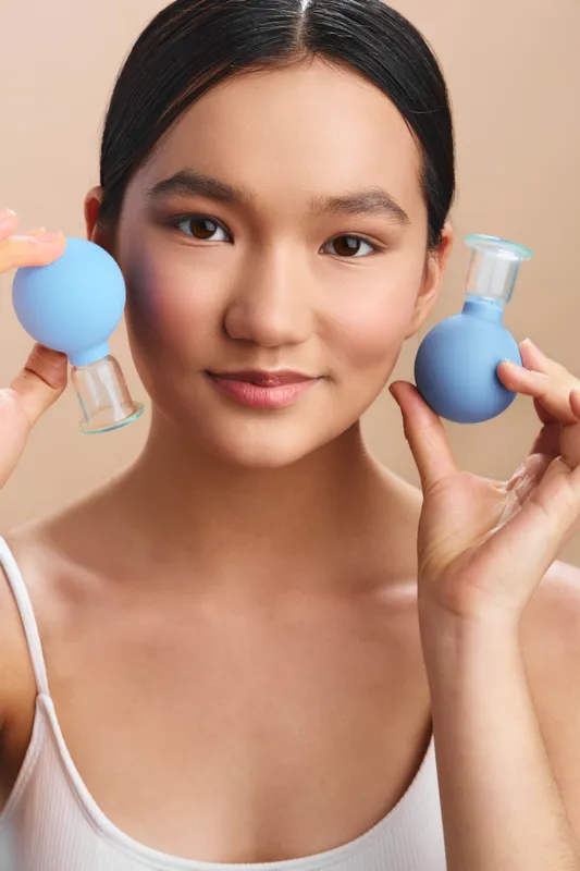 asian woman is seen performing a light workout in a calm indoor space, holding small blue dumbbells near her cheeks with a smile, conveying focus, energy, and a healthy lifestyle.