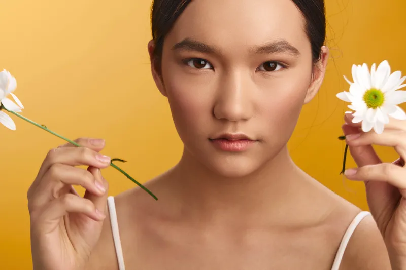 Asian woman holds daisies in both hands against a warm orange backdrop, gazing softly at the camera and projecting a serene, natural beauty with a gentle, uplifting mood.
