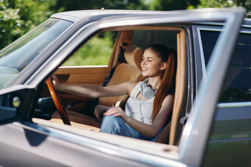 Happy young woman with long hair driving a car, wearing casual clothes and smiling in a sunny outdoor setting, capturing the essence of freedom and adventure.