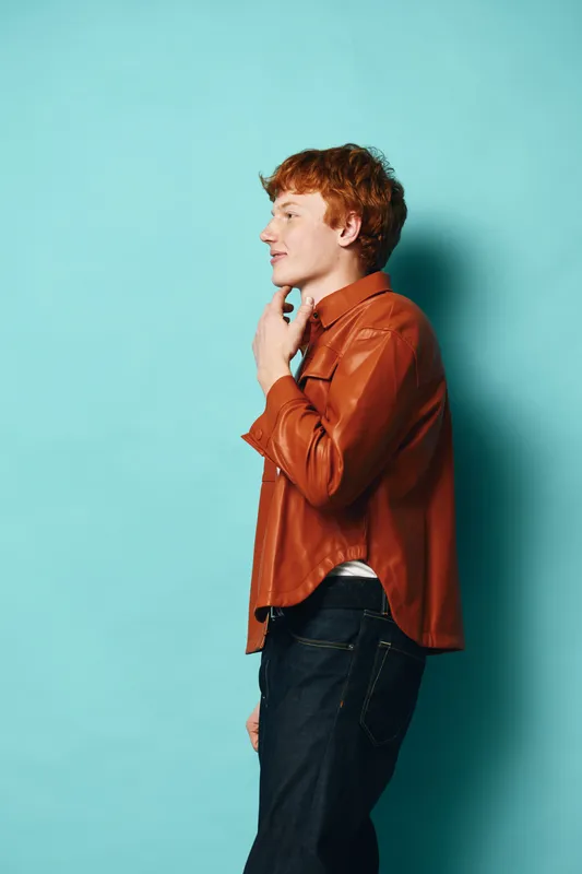 Young man with ginger hair wearing brown leather jacket and dark jeans standing in profile against blue background thoughtful and relaxed posture casual fashion style.