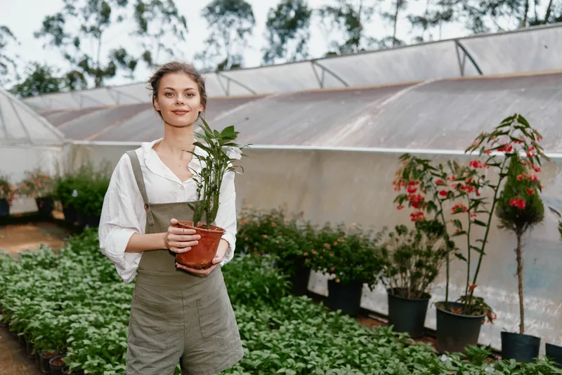 Young woman in a greenhouse holding a potted plant, wearing casual overalls and a white shirt, surrounded by greenery and flowers, showcasing a sustainable lifestyle