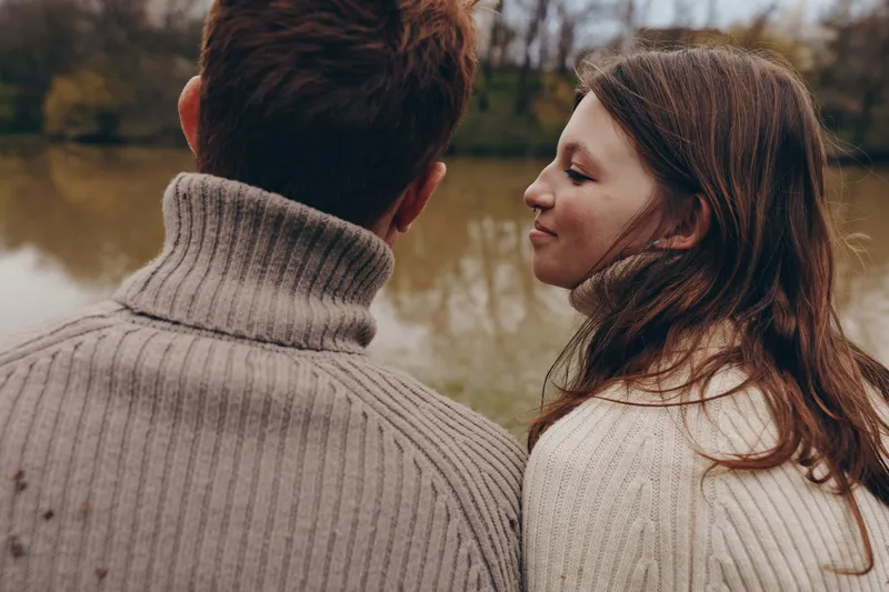 Two people in knit sweaters stand close by a pond, sharing a quiet moment outdoors. Soft natural light highlights their connection and the serene landscape. Subtle romance, calm atmosphere, and
