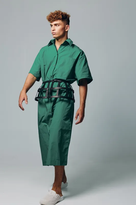 Young man with curly hair wearing green oversized shirt and pants with black belt accessory standing against gray background in studio fashion portrait.