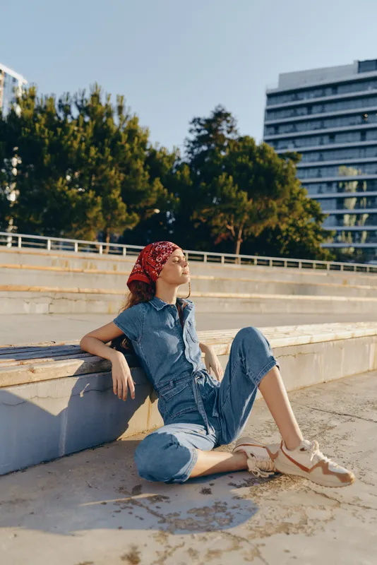 Lifestyle woman in modern boho-western denim street style relaxing outdoors, wearing a red bandana and sandals, captured with warm natural light and film color effect.
