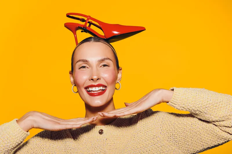 Smiling woman with red high heels on her head poses against bright yellow background. She wears beige sweater and gold hoop earrings. Concept of fun, fashion, and playful mood.