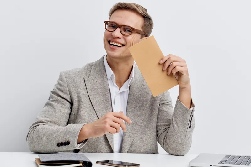 Smiling man wearing glasses and gray blazer holding a cardboard piece smiling happily in a studio setting with neutral background. Business casual professional portrait concept
