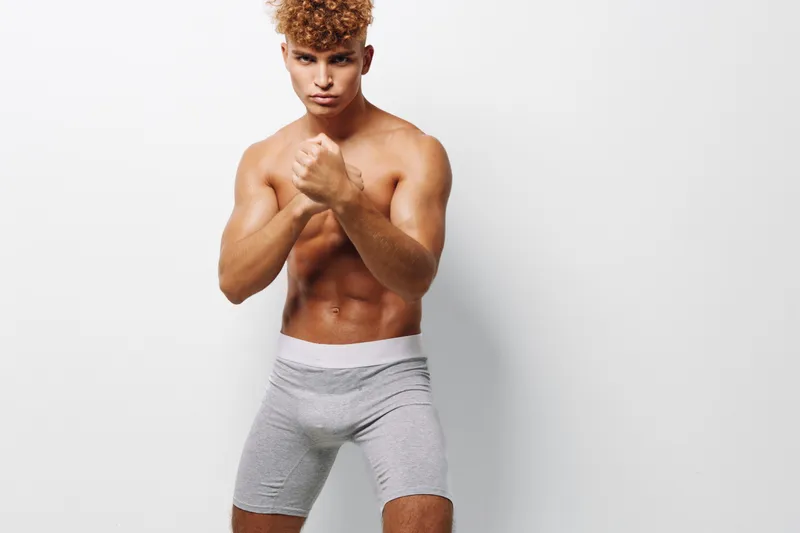Athletic young man with muscular body posing in fighting stance wearing gray shorts against white background. Fitness, strength and boxing training concept with confident expression.