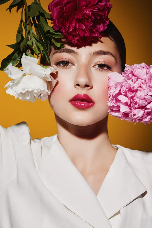 Young woman with bright fuchsia lipstick surrounded by peony flowers in white blouse on yellow background. Creative beauty and fashion shoot with floral theme.