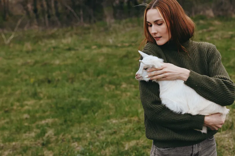 Young woman outdoors in a knitted sweater gently holds a white goat kid in a grassy field, expressing calm rural lifestyle, tender animal companionship, and peaceful connection with nature.