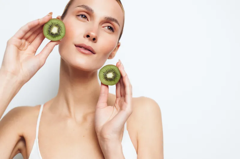 Young woman holding two halves of fresh kiwi fruit near her face on white background. Concept of healthy lifestyle, natural beauty, and fresh nutrition. Bright soft lighting.