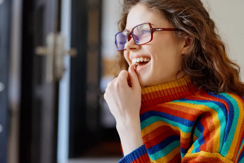 A cheerful woman in a rainbow striped sweater and glasses smiles by a window at home, a candid lifestyle moment that feels bright, warm, and relaxed.