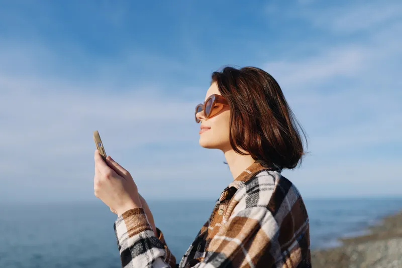 Woman, sunglasses and plaid shirt outdoors by the ocean, looking at her phone, a casual lifestyle moment with seaside fashion and a relaxed coastal vibe.