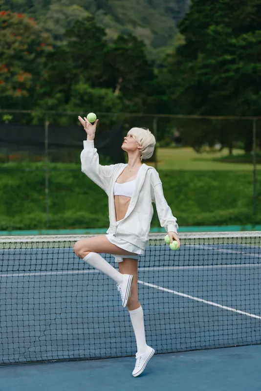 Young woman in sporty outfit showcasing a playful emotion while juggling tennis balls on a tennis court surrounded by greenery, emphasizing an active lifestyle
