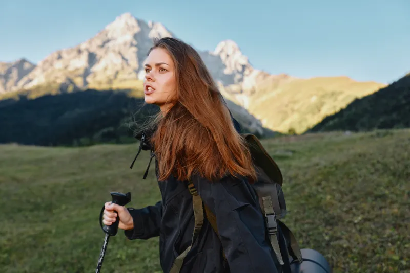 A young woman hikes across a grassy valley with a hiking pole and backpack under clear blue skies. Her windblown hair and determined expression convey adventure and exploration.