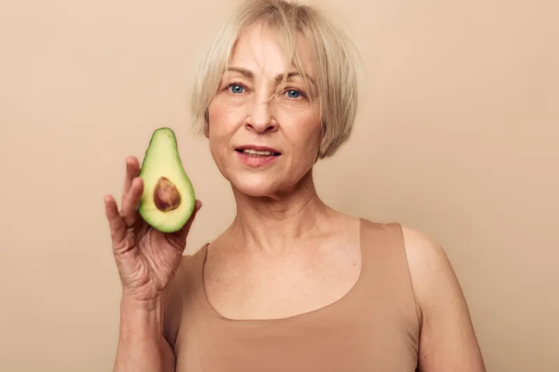 Mature woman with short blonde hair holds half of fresh avocado in hand. Neutral background and natural light highlight healthy lifestyle and organic nutrition concept.