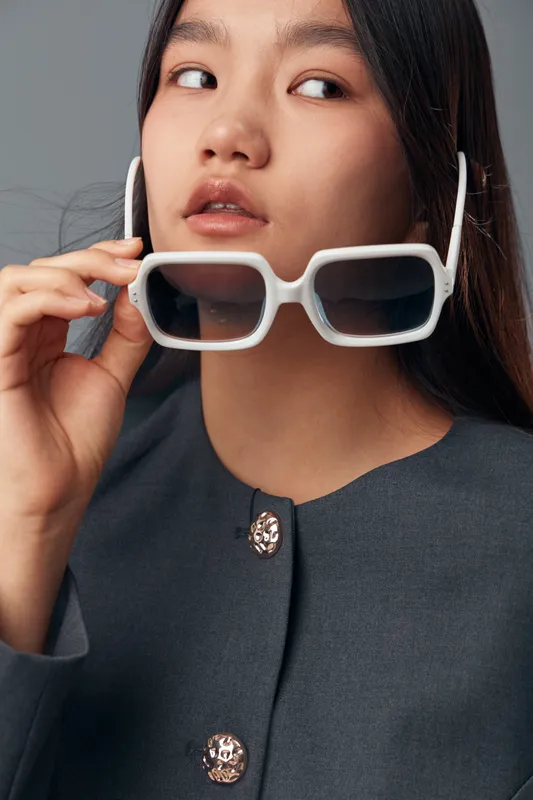 Young woman with long dark hair wearing stylish white square sunglasses and grey buttoned shirt posing against neutral background, fashion portrait closeup with natural makeup.