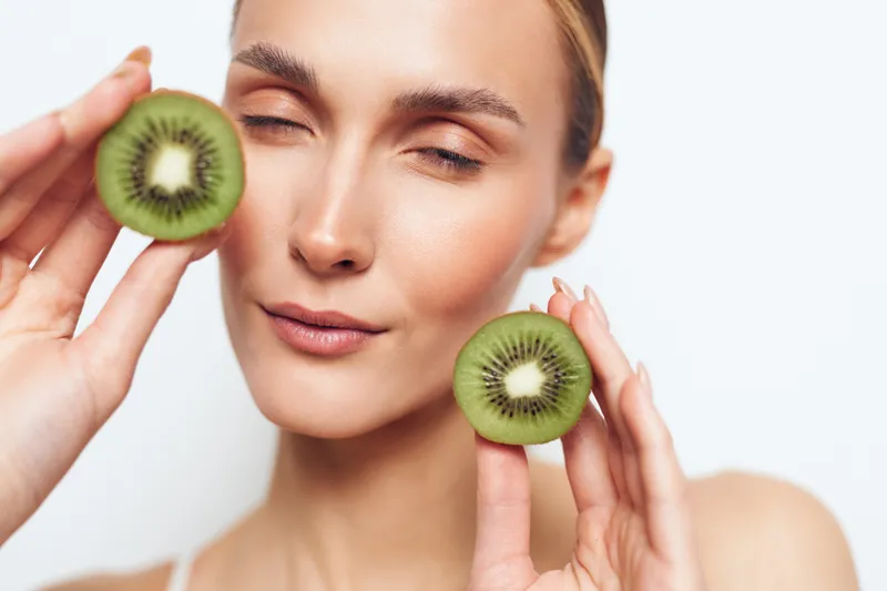 Young woman with fair skin holds kiwi slices near her cheeks, showcasing natural skincare and wellness in a soft studio setting.