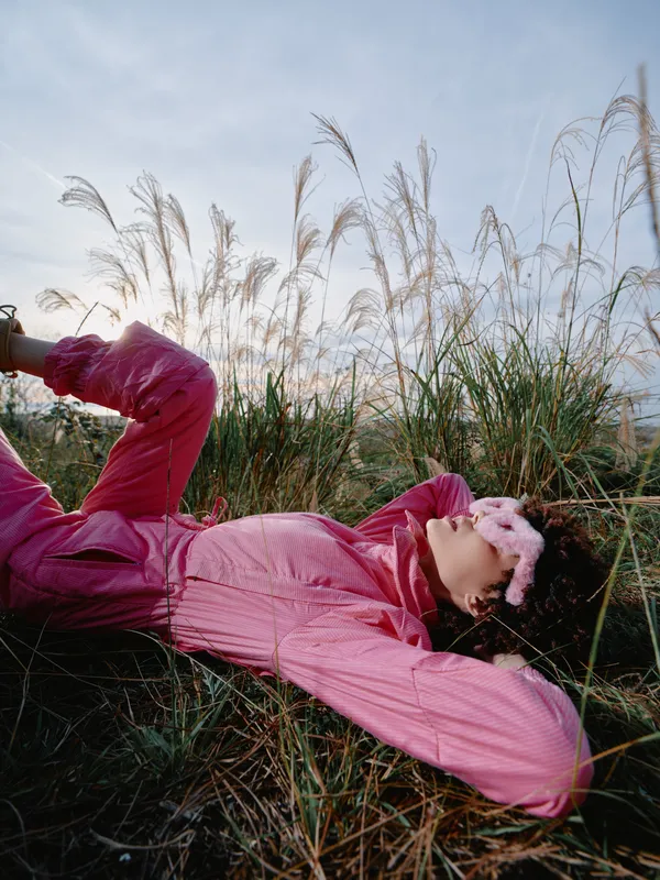 Person wearing a pink jacket lying in tall grass outdoors at sunset, relaxed pose with eyes closed, nature scene, soft light, calm mood, fashion inspired outdoor relaxation.