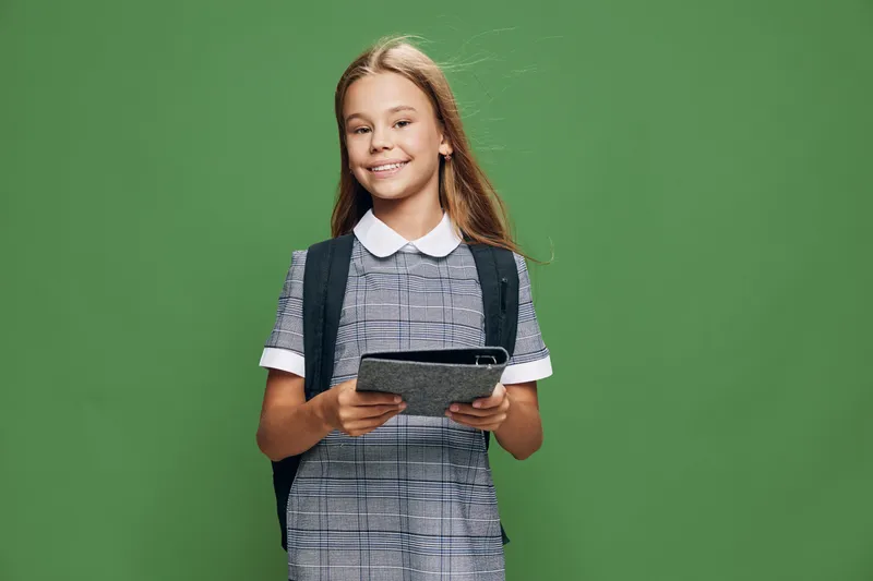 Girl student with backpack holding tablet and smiling standing against green background for education, technology and learning at school or college. Young female child with digital device.
