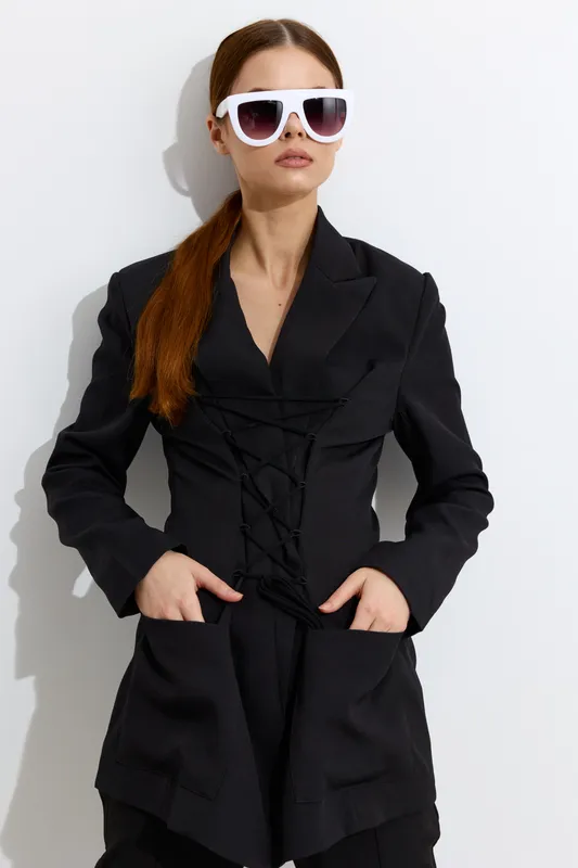 Fashion woman with long red hair wearing oversized white sunglasses and a black lace-up jacket posing confidently against a plain white background.