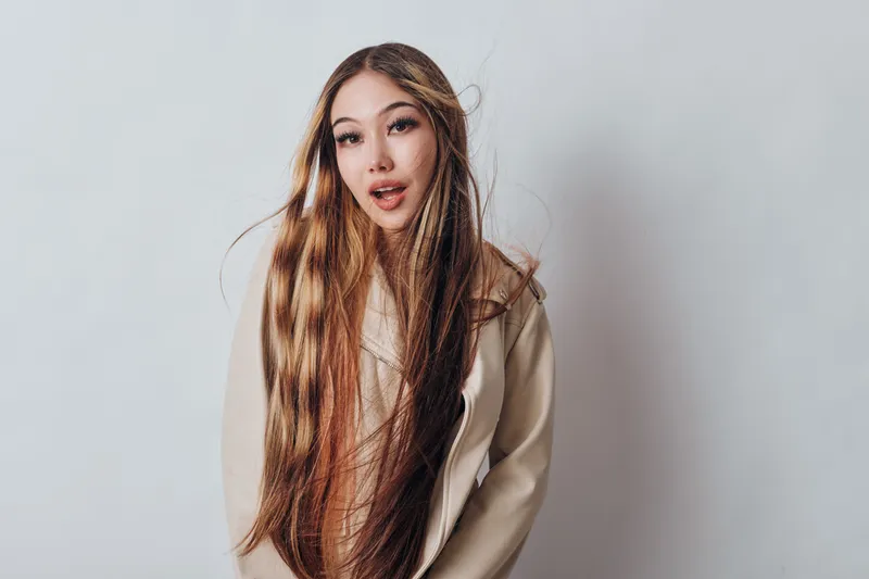 Young woman with long straight hair wearing light beige jacket posing against plain white background with natural makeup and confident expression looking at camera.