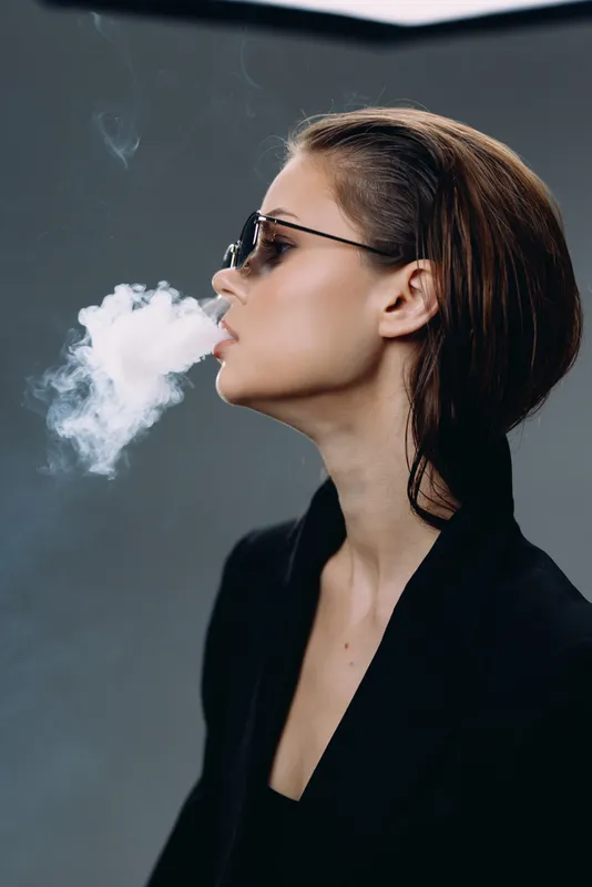 Stylish woman exhaling smoke wearing sunglasses and black blazer in studio with dark background.