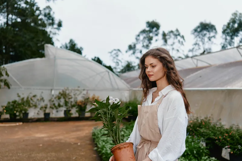 Young woman with long hair holding a potted plant in a garden, wearing an apron, exhibiting a serene expression amidst a natural, green backdrop