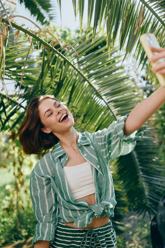Joyful woman takes a sunny selfie among lush palm leaves, wearing a striped green shirt over a white crop top, capturing a bright tropical mood.