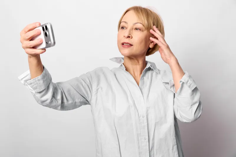 Woman takes a selfie with a smartphone, holding it near her face while touching her ear, surprised and stressed, in a bright studio with clean white background, communication concept