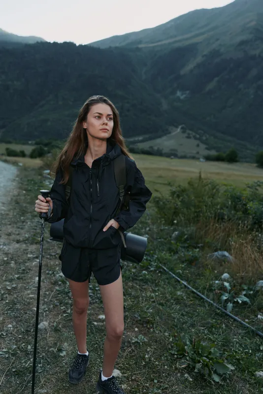 A young woman stands on a rugged trail in a mountain landscape, holding a trekking pole and camera gear. She wears a hooded jacket and shorts, gazing ahead with a calm, focused expression.