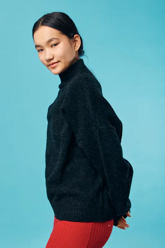 Young woman posing in black sweater and red skirt against blue background with relaxed expression and casual style. Female model with dark hair and minimal makeup in studio portrait.