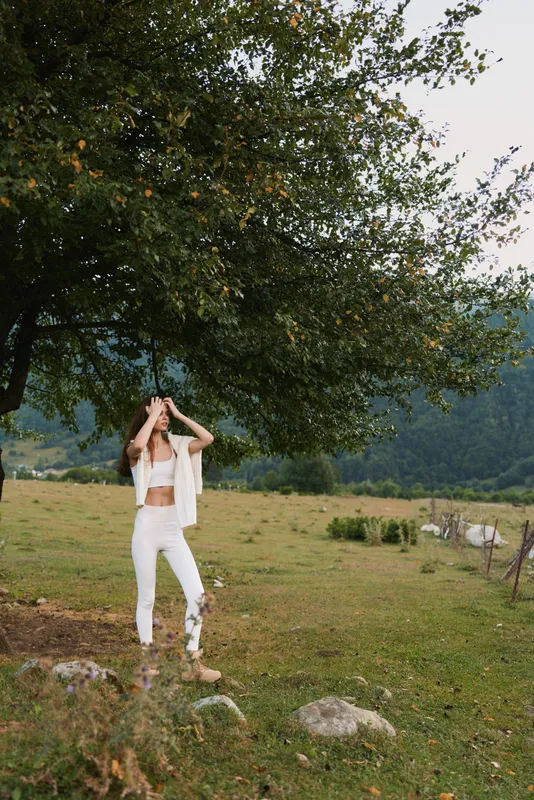Young woman stands in an open outdoor field beneath a leafy tree, wearing a white outfit and relaxed pose, bathed in warm natural light and fresh summer air.