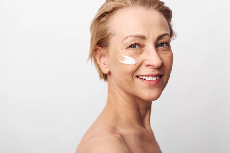 Smiling middle aged woman with short blonde hair applies white face cream on cheek. Close up on light background. Concept of skincare, beauty routine, self care, and healthy skin glow.