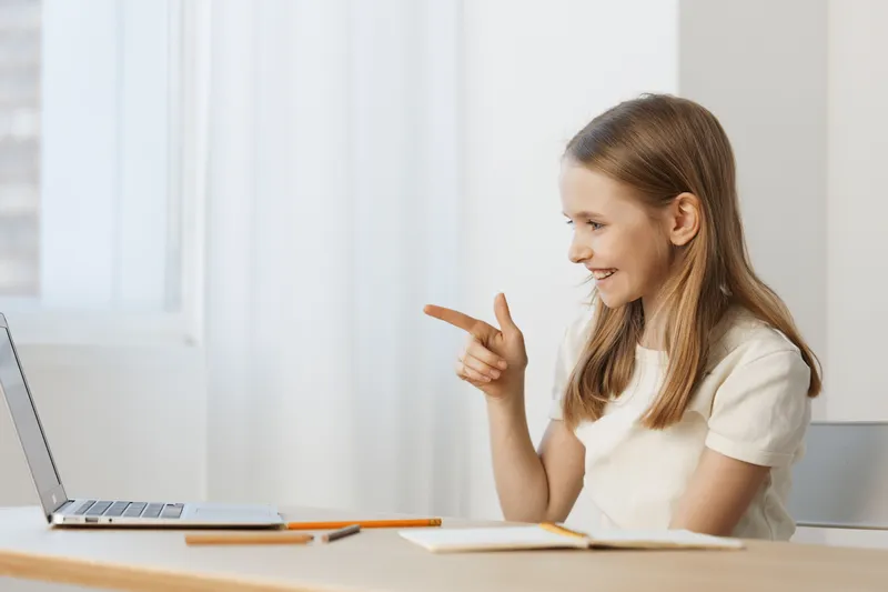 Young girl engaging with a laptop, showing excitement and joy, wearing a casual white shirt, in a bright room with a minimalistic design
