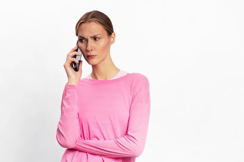Concerned woman in pink sweater talks on phone looking away with serious expression isolated on white background. Concept for communication, stress, and emotional response.