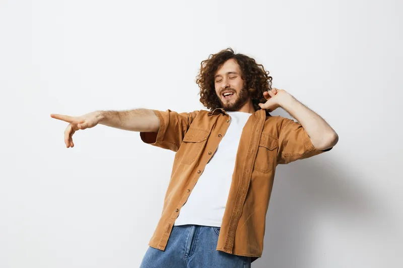 Young man with curly hair joyfully dancing against a plain background, wearing a casual brown shirt and white t shirt, showcasing happiness and carefree attitude