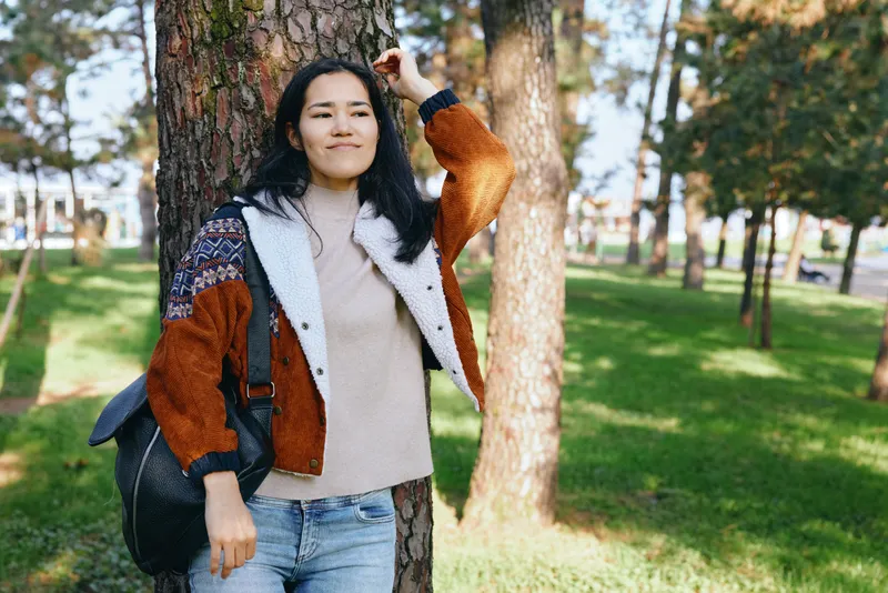 Woman in a cozy sweater and color block jacket with backpack walking through a sunlit park, enjoying nature, outdoor leisure, and autumn atmosphere