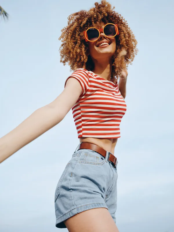 A young woman with curly hair wears a red striped crop top and denim shorts, posing cheerfully under a sunny blue sky, exuding carefree summer style.