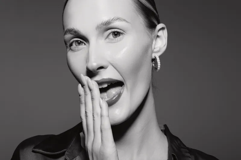 Close-up of a fair skinned Caucasian woman with slicked back hair whispering with hand near mouth. Studio portrait in black and white with soft lighting and expressive mood.