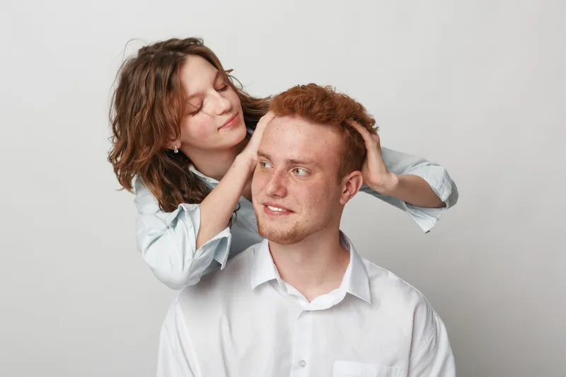 Young woman gently massages young man’s head and hair in white studio. Concept of care, relaxation, friendship, and personal connection between two people with soft natural expressions.