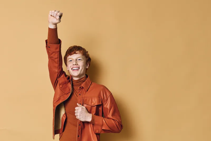 Happy young man in orange jacket smiling and raising fist in celebration on beige background. Portrait of confident male with casual autumn outfit and joyful expression.