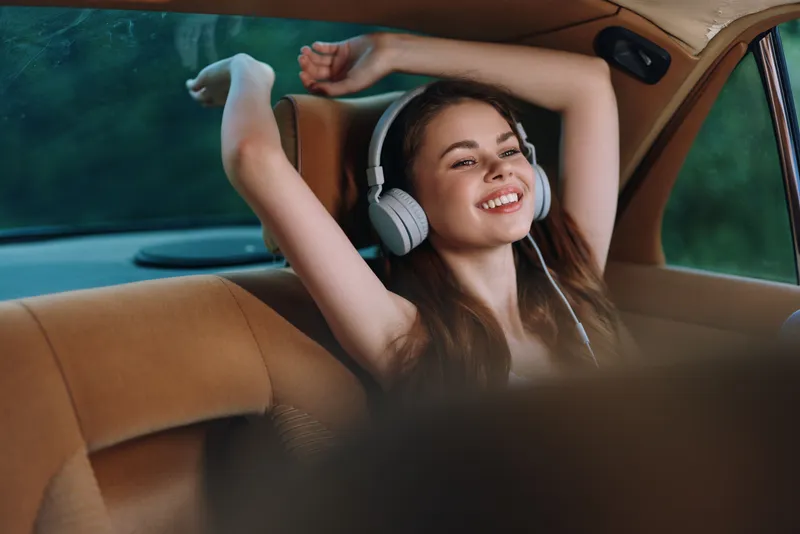 Happy young woman enjoying music in a car, showcasing joy and relaxation, surrounded by a serene forest background and a soft color palette.