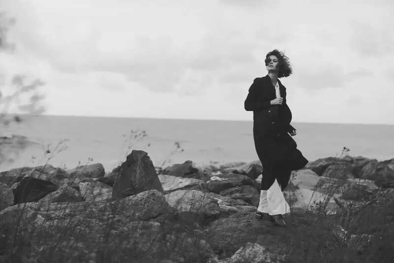 Coastline landscape fashion wind movement coat woman by the sea among rocks, monochrome mood, shoreline solitude, outdoor drama, graceful silhouette and flowing garment capturing style, mood, and