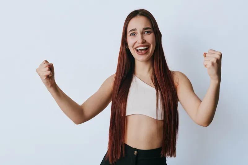 Young woman with long hair and crop top smiling and showing fists in victory gesture on plain background. Female person expressing happiness and success with confident pose.
