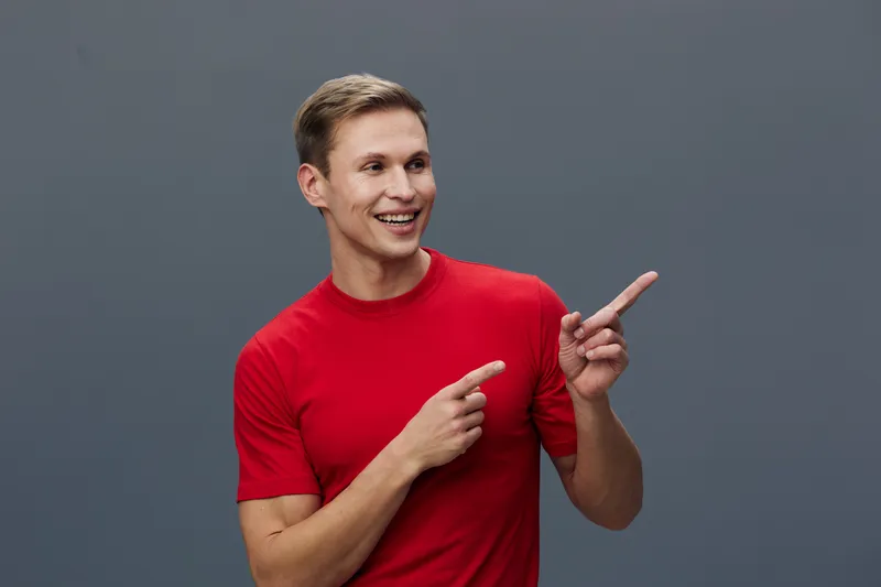 Smiling man in red t-shirt pointing with finger isolated on gray background digital nomad concept happy young caucasian man lifestyle