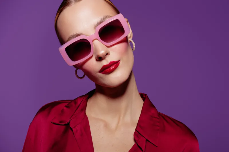 Stylish woman with pink sunglasses and red lipstick poses against purple background wearing red blouse and hoop earrings. Concept of fashion, beauty, and confident attitude.