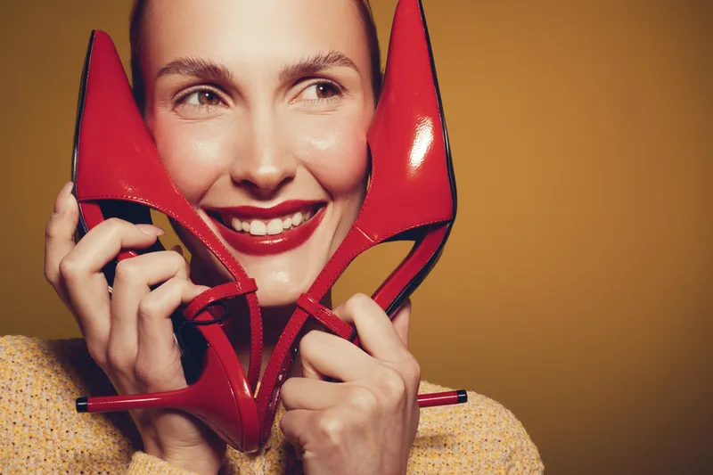 Smiling woman holds red high heel shoes near face, showing bright lipstick and joyful expression. Concept of fashion, beauty, footwear, and happiness with warm studio lighting.